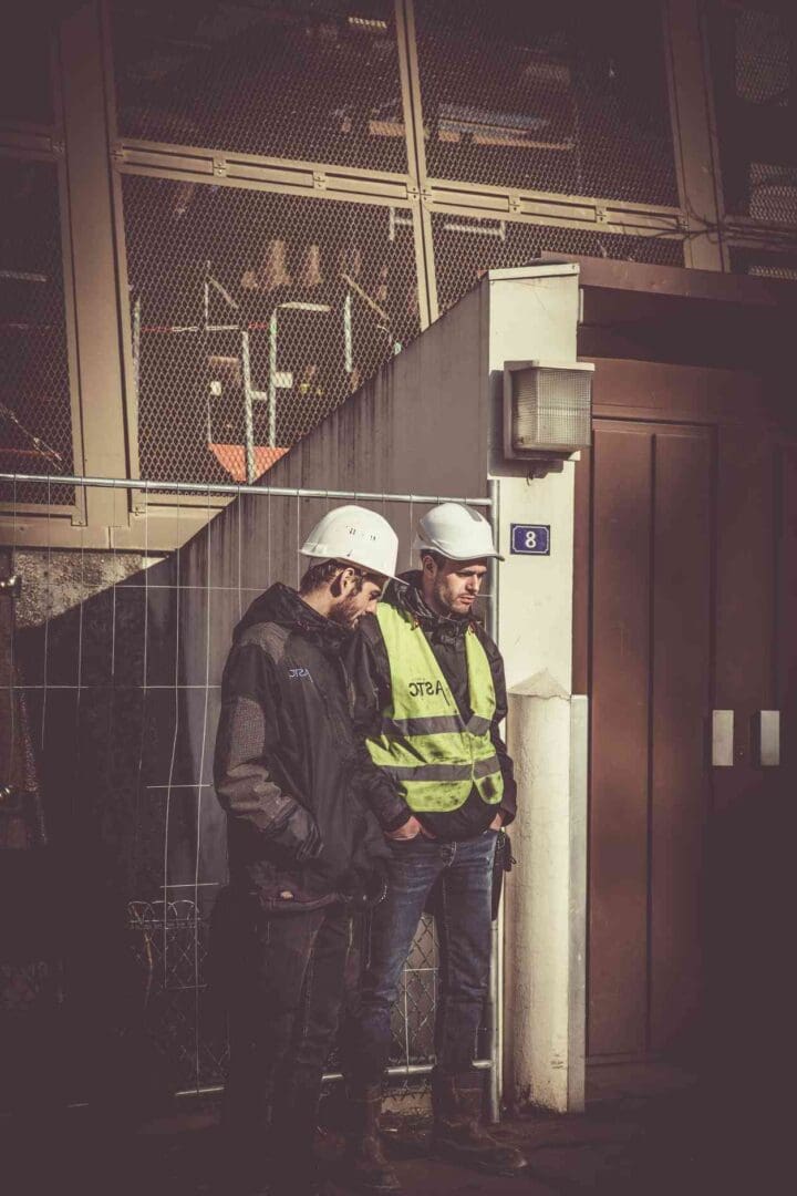 Two men in hard hats and jackets standing next to a building.