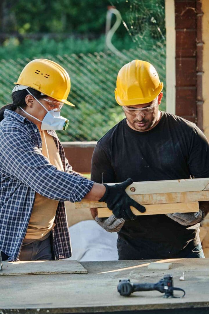 Two people wearing hard hats and gloves holding a wooden board.