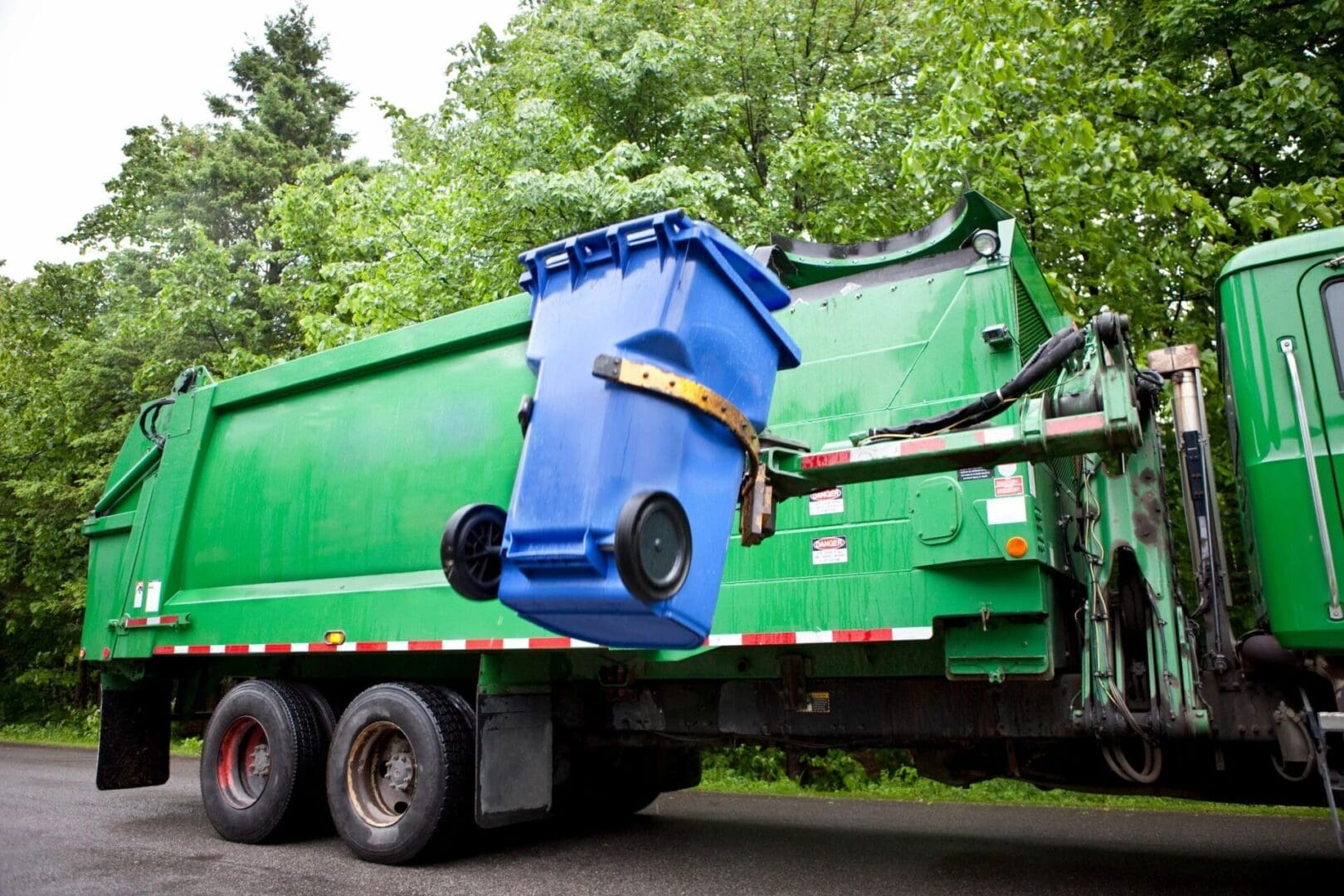 A garbage truck with a blue trash can on the back.